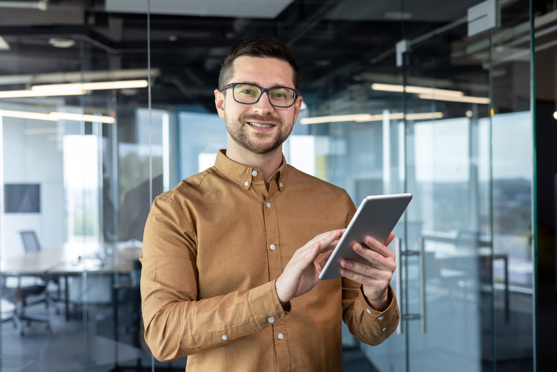 Portrait of a young male designer standing in the office and using a tablet. He looks at the camera with a smile