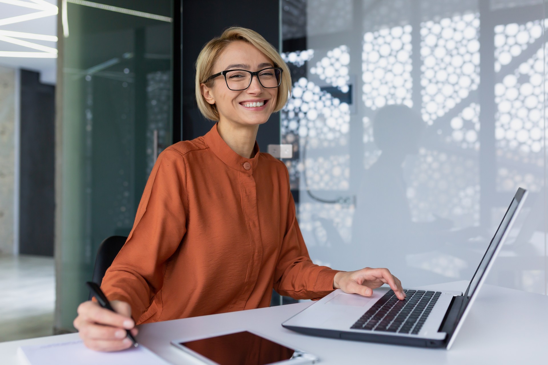 Successful and happy business woman looking at camera sitting inside office working with documents, portrait of satisfied woman writing in notebook and working using laptop at work