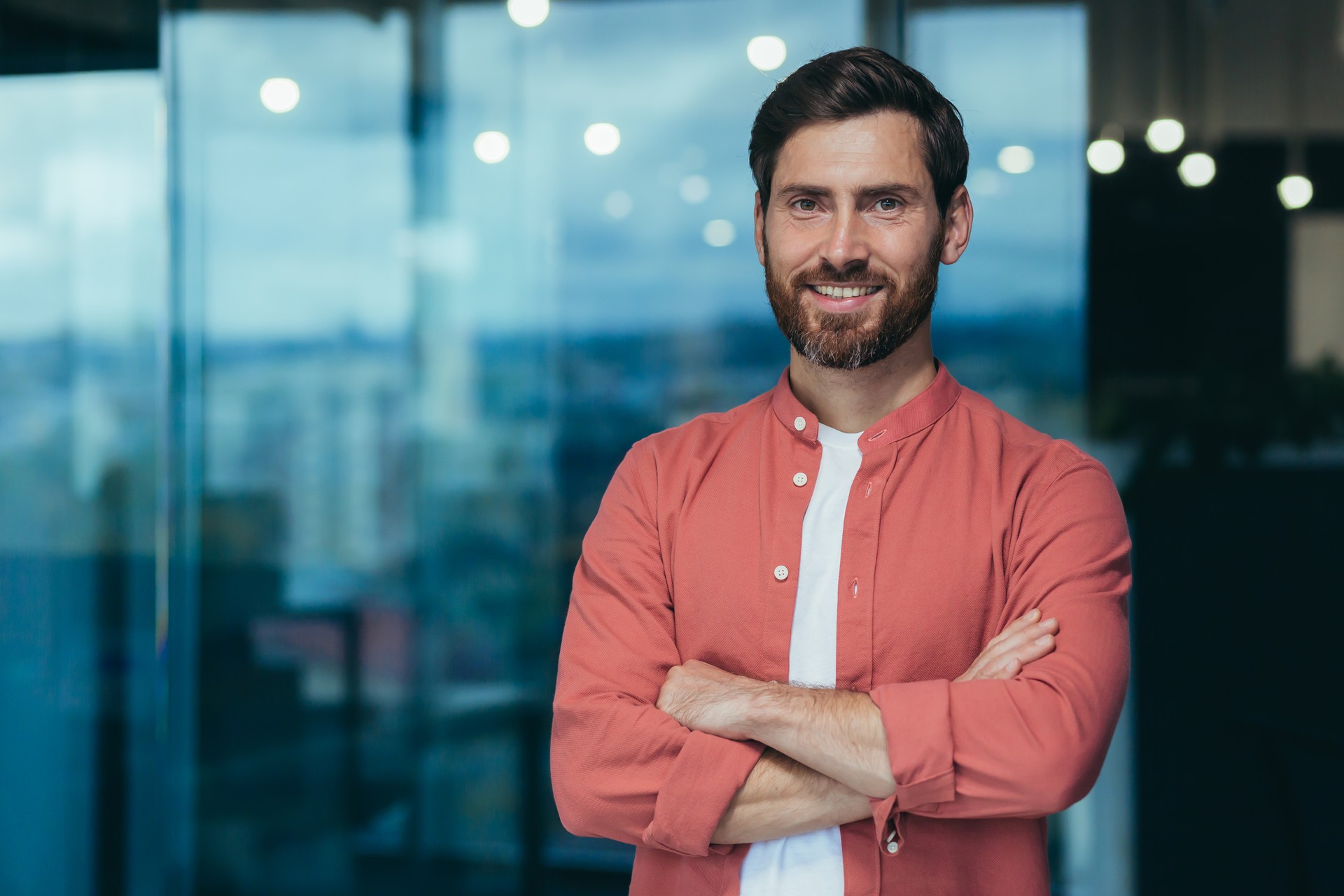 Portrait of a happy and smiling programmer in the middle of a modern office, a man in glasses and a red shirt is looking at the camera with his arms crossed